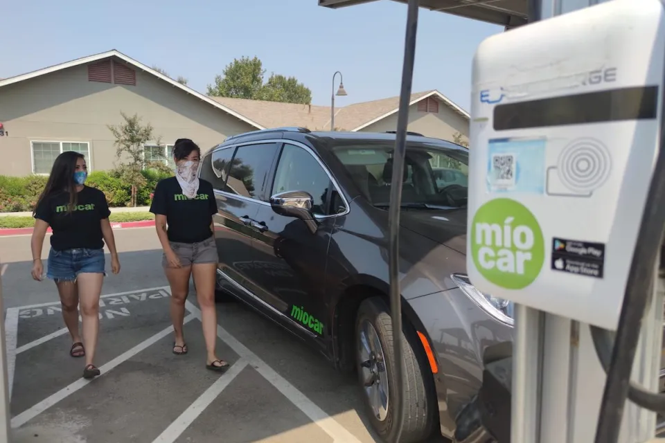 Black SUV charging at Miocar electric car sharing station with two women walking towards it.
